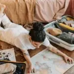 A woman lying on the floor indoors, planning a trip with a map and open suitcase.